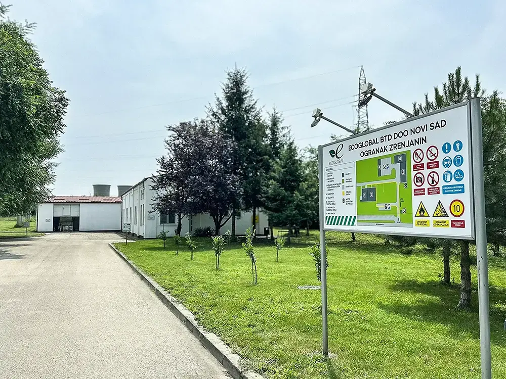 An outdoor scene with a paved pathway leading towards a building complex under a clear sky. On the right, there is a large informational signboard with various symbols and text, predominantly in green and yellow colors. The signboard includes illustrations of industrial processes or services offered by the facility. Surrounding the pathway are neatly planted young trees and well-maintained grass, with power lines visible in the background