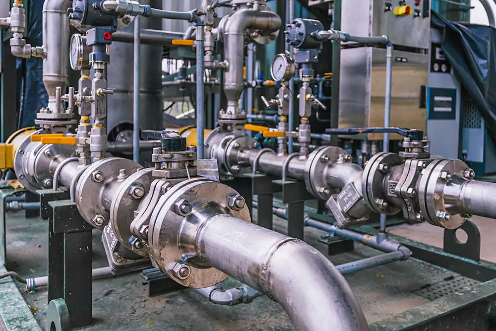 An industrial setting with a series of metal pipes and valves. The pipes are interconnected with flanges and bolts, featuring various gauges and handwheels. The equipment appears well-maintained, suggesting a controlled environment possibly for manufacturing or processing purposes. The complexity of the piping system indicates its role in a facility that requires precise control of fluid movement.