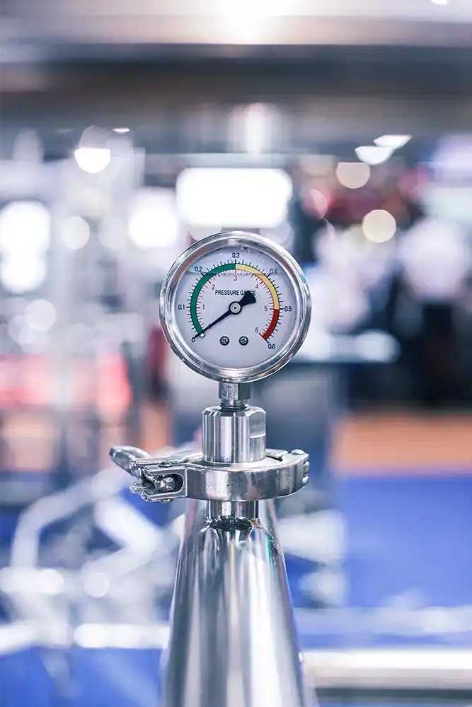 A close-up of a pressure gauge mounted on top of a metal cylinder, with readings in the green zone indicating safe pressure levels. The background is blurred with hints of gym equipment, suggesting the setting is a fitness center or similar environment where the cylinder might be used for equipment maintenance or operation.