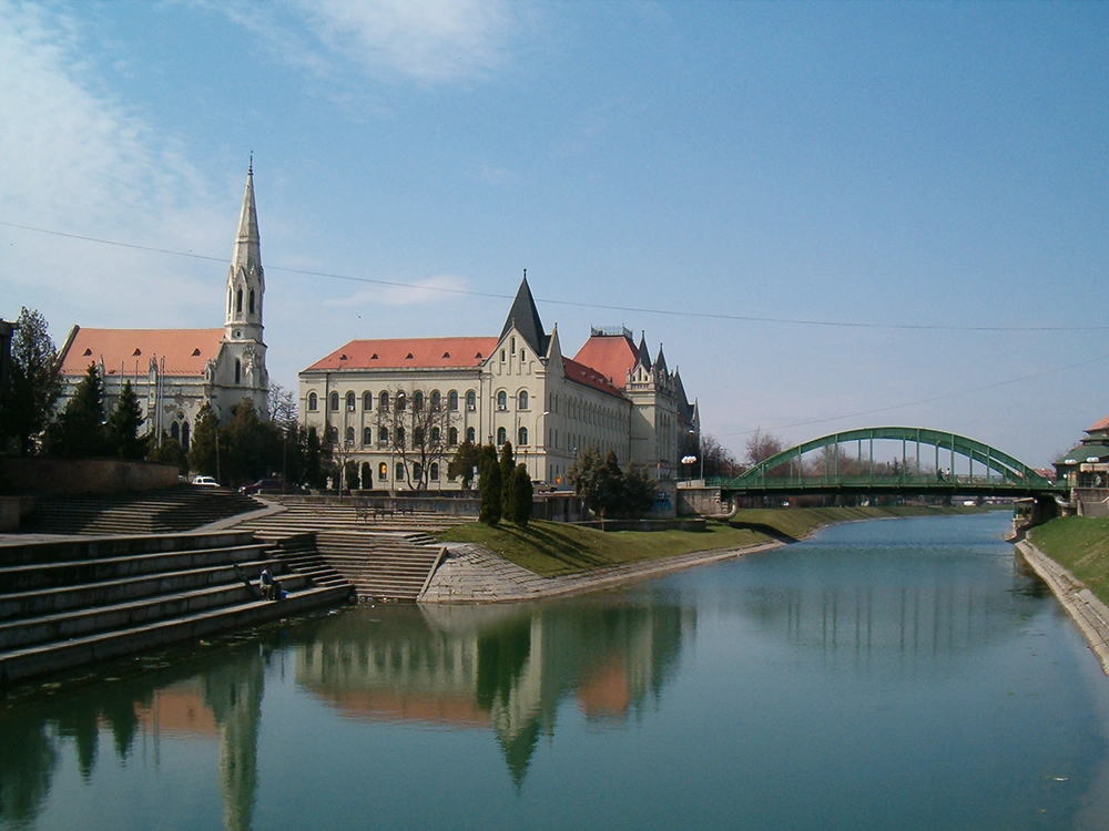 The image shows a serene river scene with clear blue skies above. On the left, there is a grand white building with a prominent spire and multiple arched windows, resembling a church or cathedral. Adjacent to it is another large building with similar architectural features. In the foreground, there are steps leading down to the riverbank. A green arched bridge spans across the river in the background. The calm water reflects the buildings and bridge, creating a symmetrical effect that adds to the tranquility of the scene.